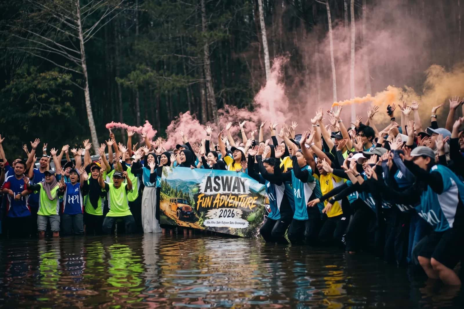 Sekelompok besar orang berkumpul di air dangkal di depan hutan pinus, merayakan "ASWAY FUN ADVENTURE 2026". Mereka memegang spanduk besar, bersorak dengan tangan di atas, dan bom asap merah muda dan oranye menyala di sekitar mereka.